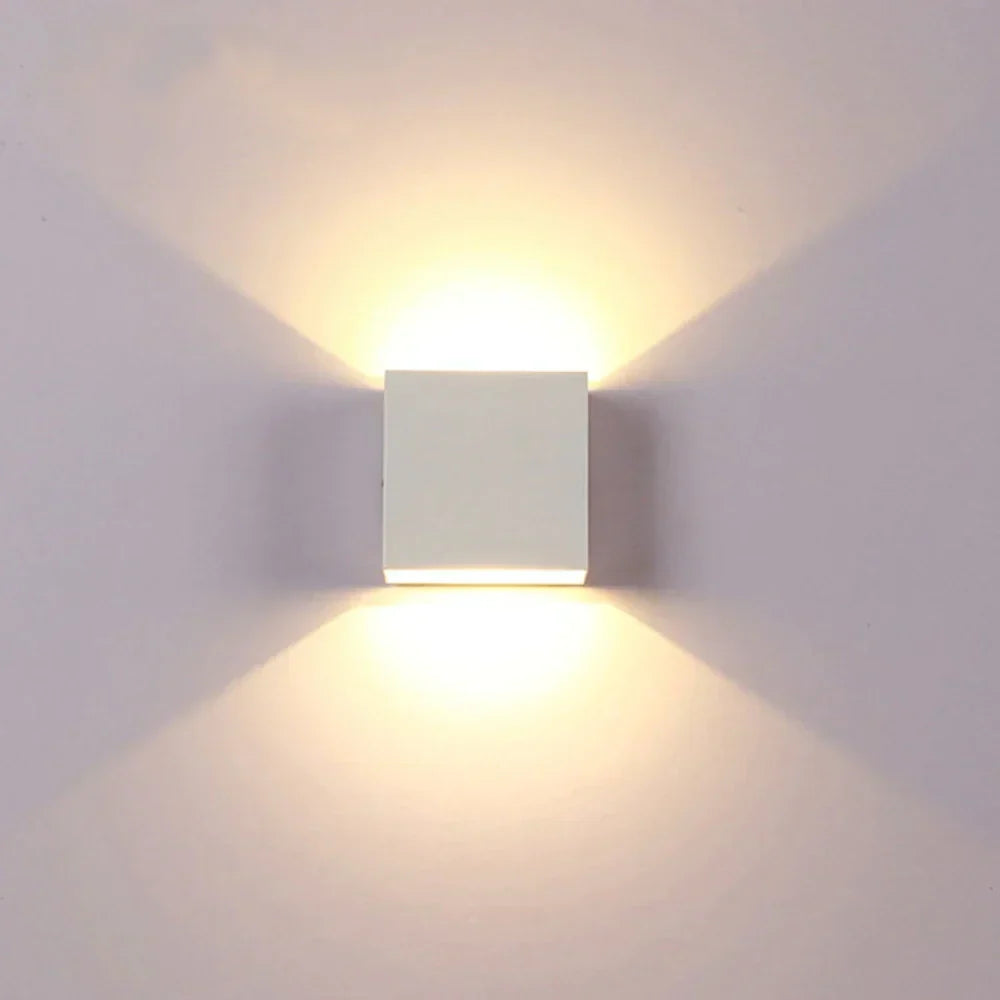 Front view of a white cube wall lamp glowing warmly, with light fanning out above and below on a pale wall. 17