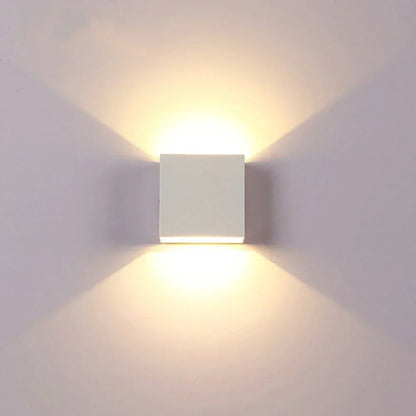 Front view of a white cube wall lamp glowing warmly, with light fanning out above and below on a pale wall. 17