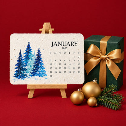 January card with blue watercolour fir trees on a wooden easel beside a green gift and gold baubles 2