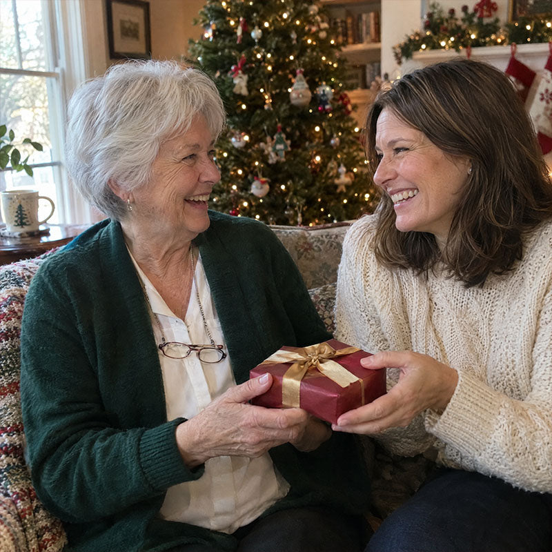 Two smiling women on a sofa by a Christmas tree, one handing the other a wrapped gift 4