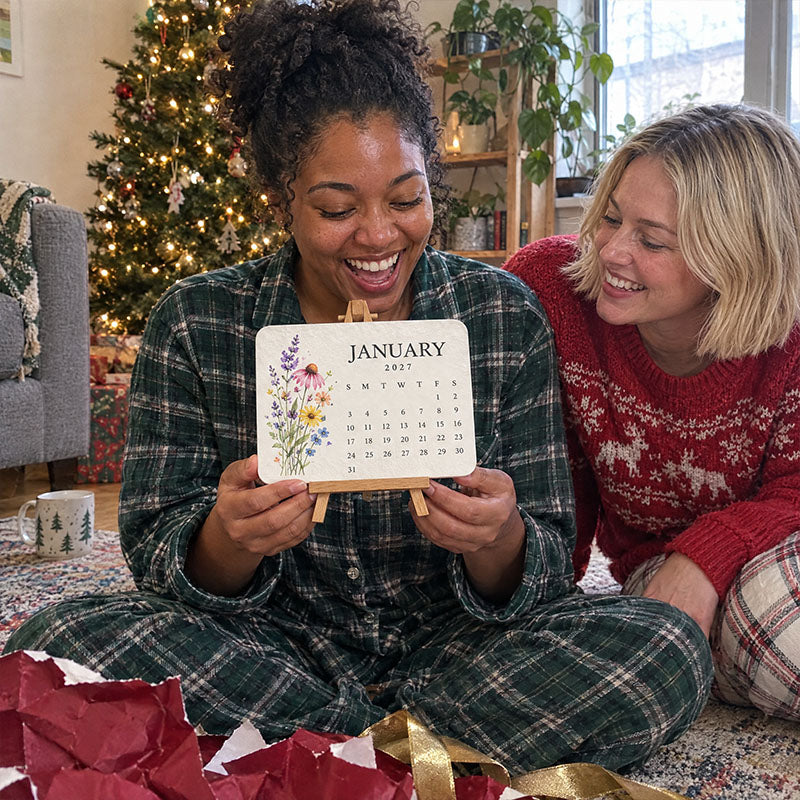 Laughing woman in tartan pyjamas holds up the January wildflower card on its easel among torn wrapping paper 5