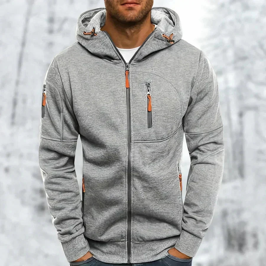 Model wearing the hoodie in light grey with a textured jacquard finish and orange zip pulls, snowy backdrop. 8