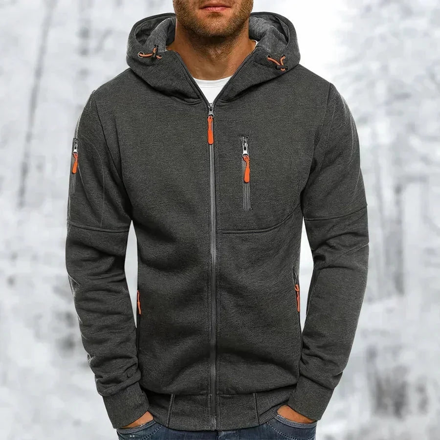 Model wearing the hoodie in charcoal grey with orange zip pulls and ribbed cuffs, hands in pockets, snowy backdrop. 9