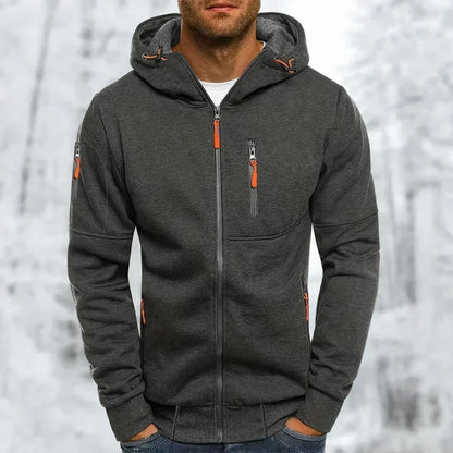 Model wearing the hoodie in charcoal grey with orange zip pulls and ribbed cuffs, hands in pockets, snowy backdrop. 9