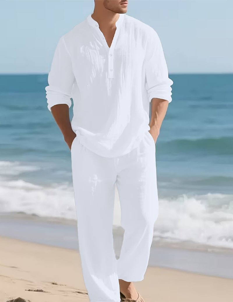 Model wearing the white set on a beach, hands in pockets, with sea and gentle waves behind. 2