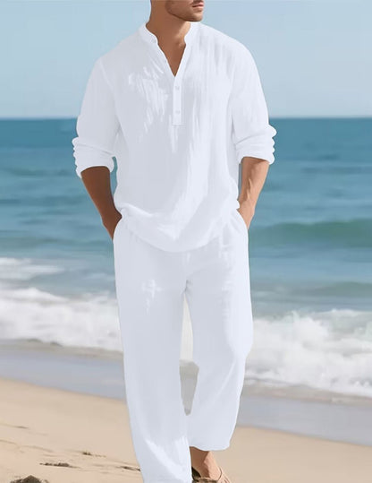 Model wearing the white set on a beach, hands in pockets, with sea and gentle waves behind. 2