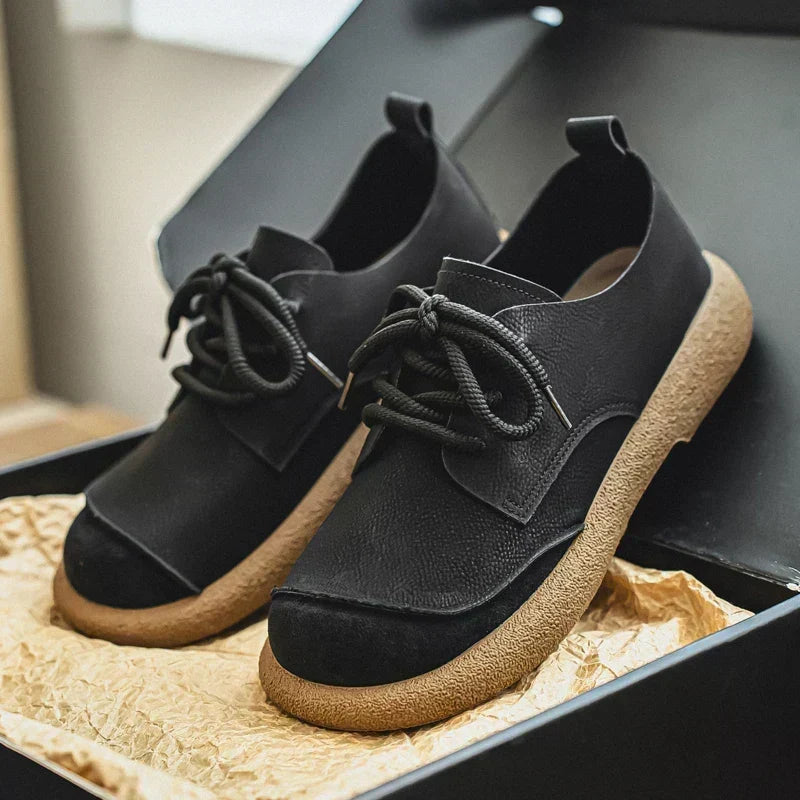 Black leather lace-up shoes with a black suede toe cap and thick tan crepe soles, displayed in an open box on crinkled paper. 12