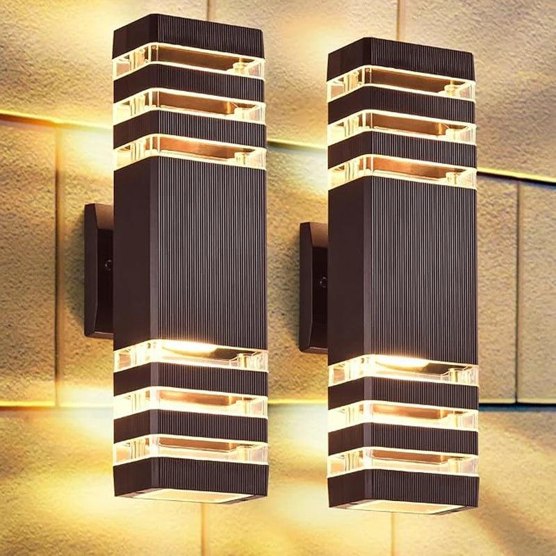 Pair of dark ribbed square wall lights mounted on a stone wall, casting warm light upwards and downwards through clear striped panels. 1