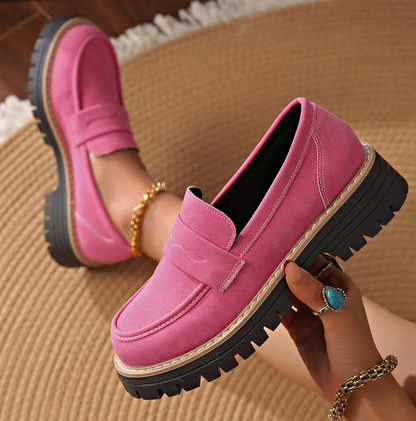 Bright pink suede-effect loafer held in a hand, with a second worn on a foot, on a woven rug. 18