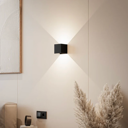 Black cube wall lamp casting light up and down on a cream wall beside pampas grass and a framed textured artwork. 1