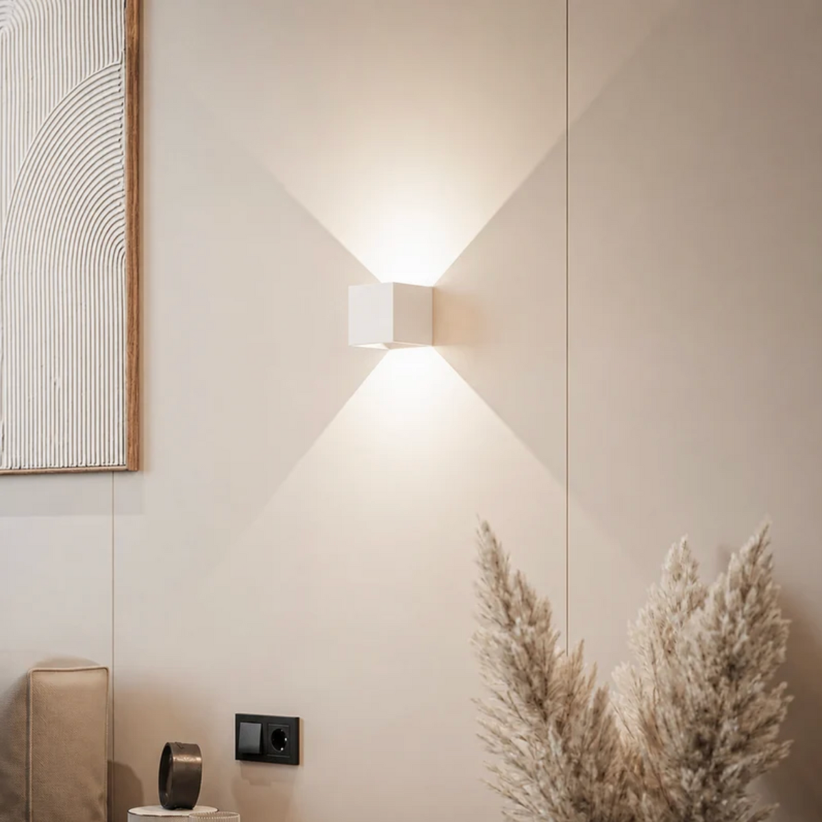 White cube wall lamp casting light up and down on a cream wall beside pampas grass and a framed textured artwork. 12