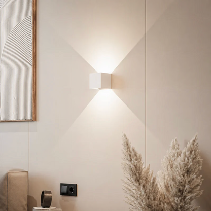 White cube wall lamp casting light up and down on a cream wall beside pampas grass and a framed textured artwork. 12
