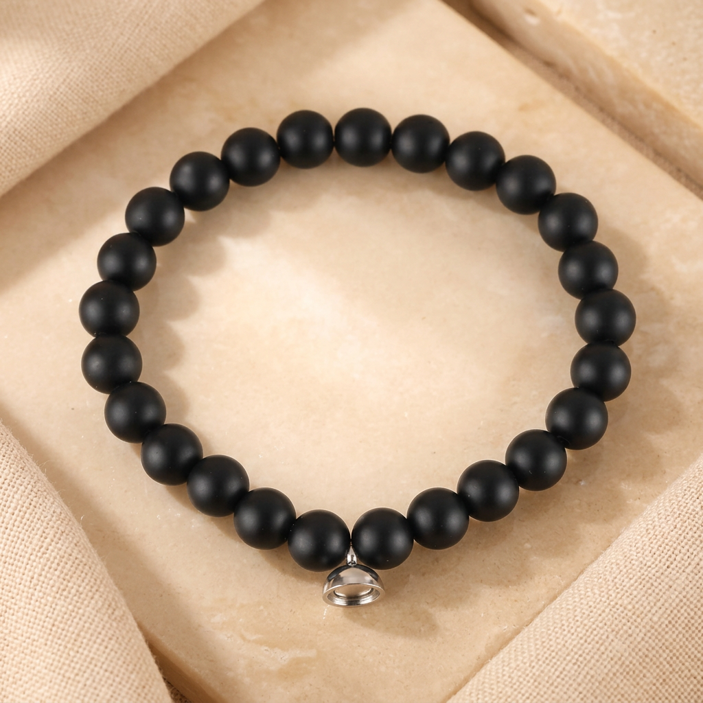 Matte black round bead bracelet with a small silver-tone magnetic charm, laid flat on a pale stone tile 2