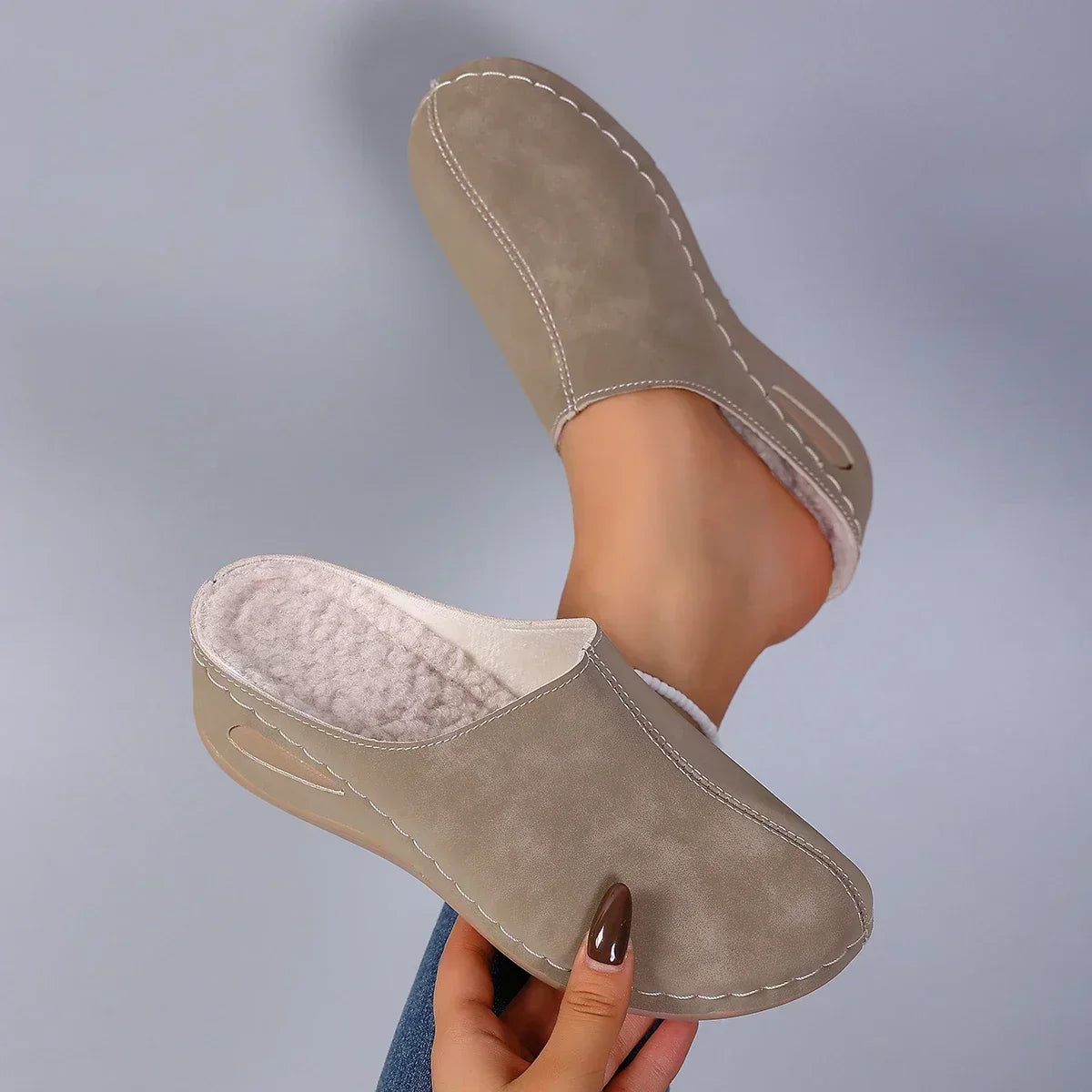 Taupe backless slippers held up by hand, showing the soft fleece interior, contrast stitching and cut-out wedge sole beside a worn slipper. 6