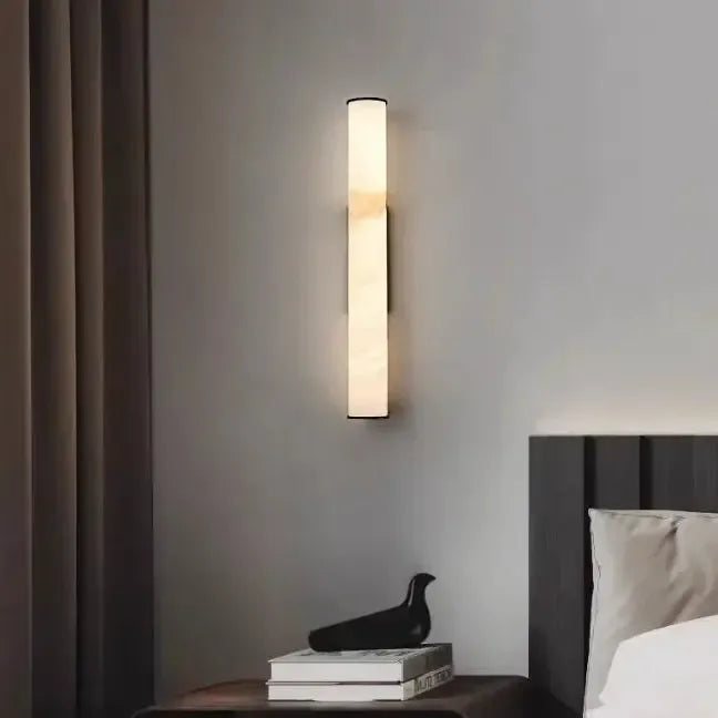 Slim marble wall light glowing above a bedside table with books and a small black bird ornament, next to a dark headboard. 14