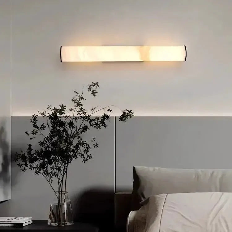 Marble wall light mounted horizontally above a bed, with a glass vase of dark leafy stems on a bedside table. 16