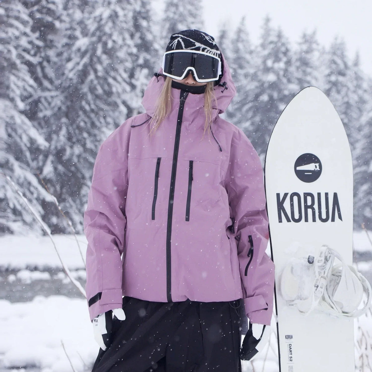 Model in the dusty pink hooded jacket stands facing the camera beside a snowboard, showing the black chest zips. 3