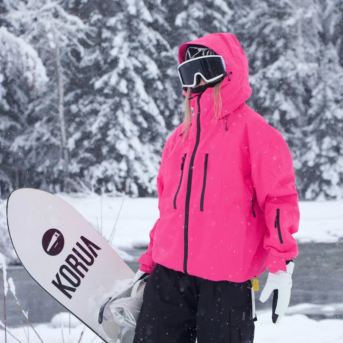 Model in the bright pink hooded jacket with the hood up, holding a snowboard in a snowy forest. 5
