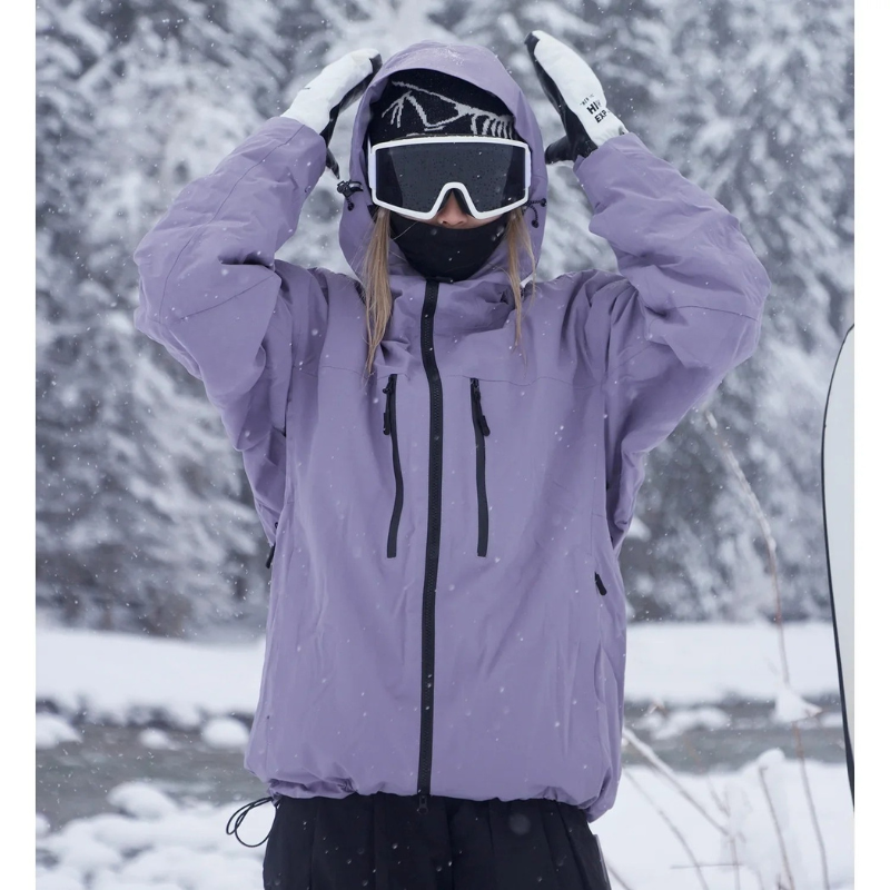 Model in a lilac hooded jacket with black zips raises both gloved hands to the hood in falling snow. 1