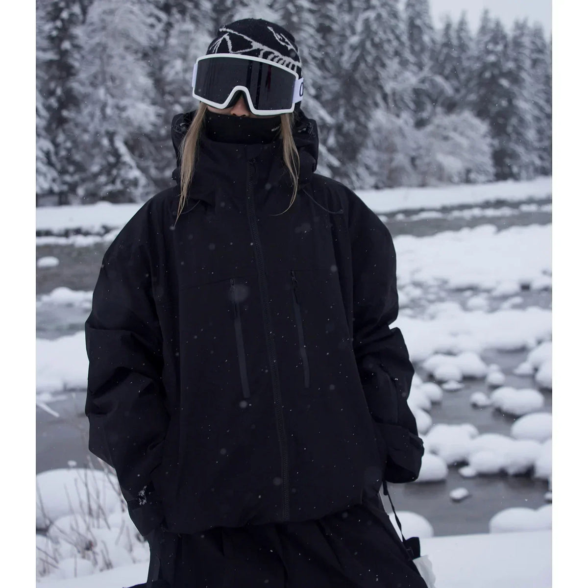 Model in the black version of the oversized hooded jacket, hands in pockets, beside a snowy river. 2