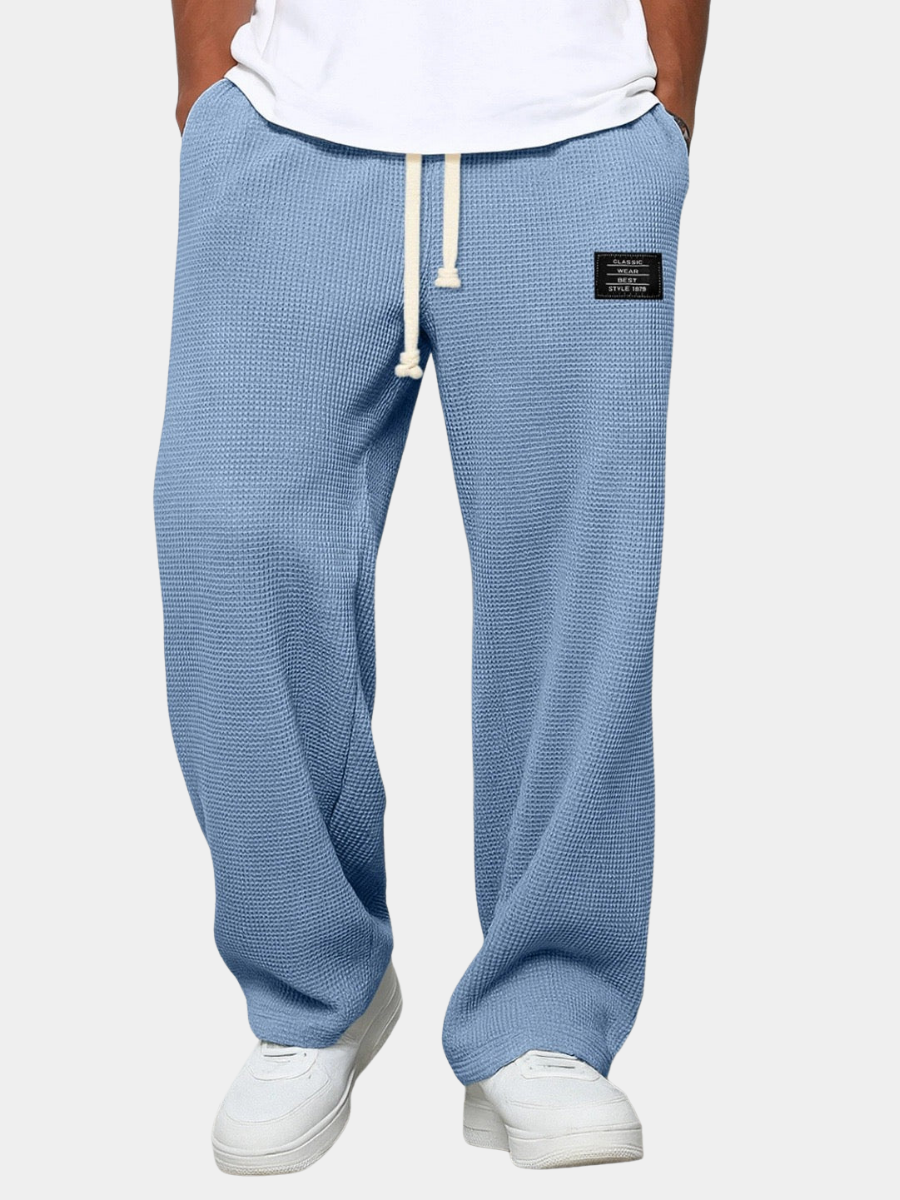 Model wearing the trousers in pale blue against a plain light background, front view, drawstring tied, hands in pockets, white t-shirt. 4