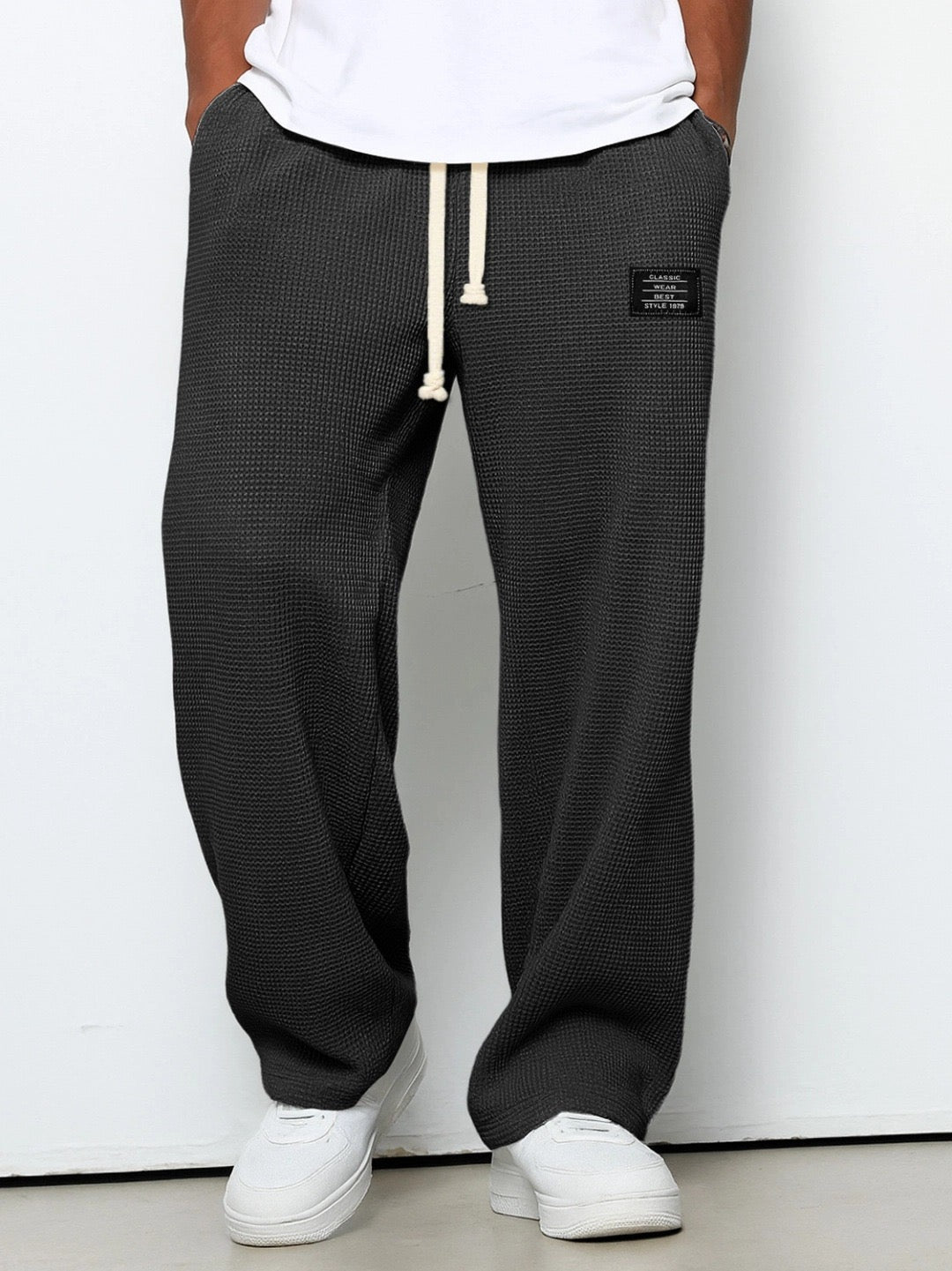 Model wearing the trousers in dark charcoal grey, front view, with cream drawstring, hands in pockets, white t-shirt and trainers. 1