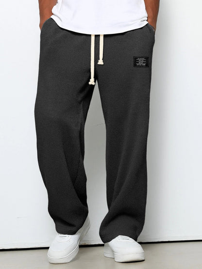 Model wearing the trousers in dark charcoal grey, front view, with cream drawstring, hands in pockets, white t-shirt and trainers. 1