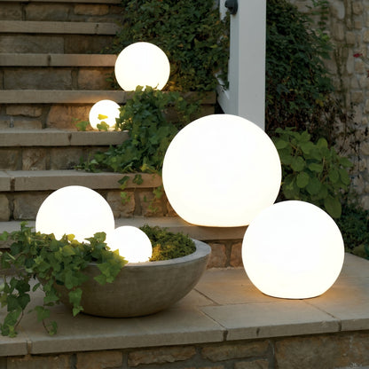 Glowing white spheres in several sizes lit at dusk on stone steps, with one resting inside a concrete planter among ivy. 1