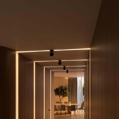 Four black ceiling lights in a wood-panelled corridor casting warm horizontal beams, with a dining table and olive tree beyond. 14
