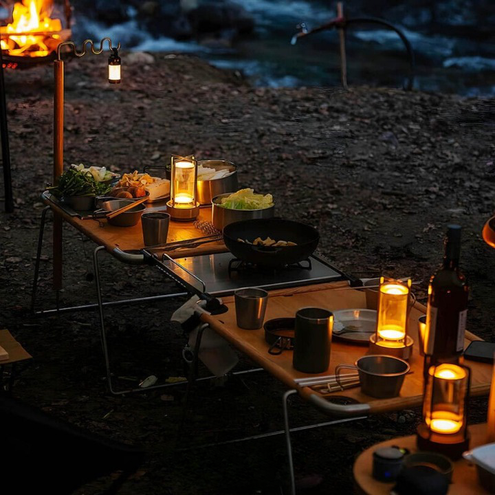 Lit lamps on folding wooden camping tables at dusk, among cookware and food, with a campfire and stream behind. 4