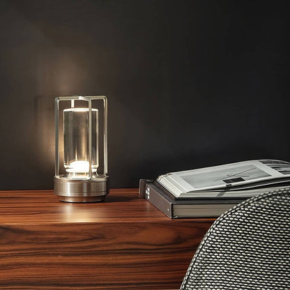 Silver-finish lamp softly lit on a polished wooden sideboard against a dark wall, next to open books and a mesh chair. 5