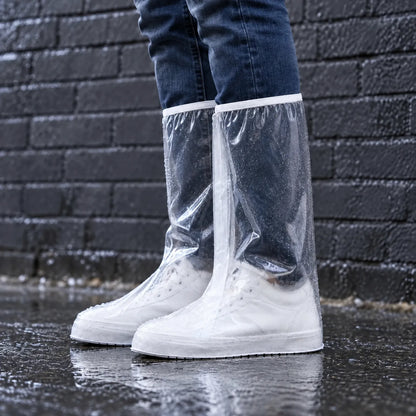 Side view of tall clear overshoes worn over white trainers and jeans, beaded with raindrops on a wet pavement by a dark brick wall. 1