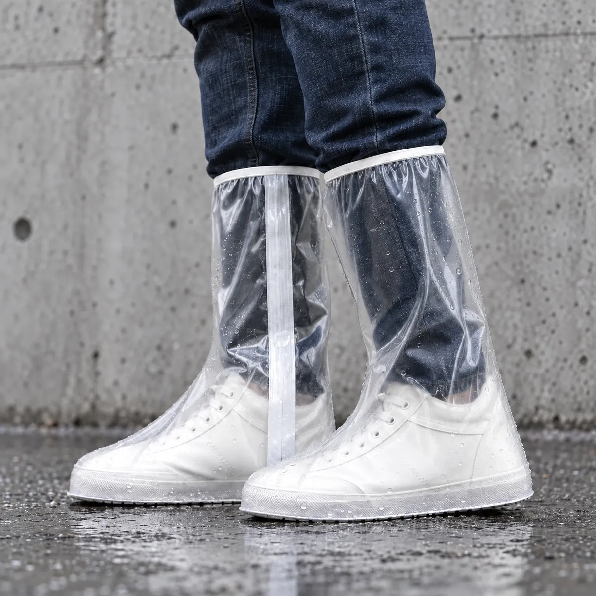 Clear knee-high overshoes with a white zip strip and raindrops, worn over white trainers against a grey concrete wall on wet ground. 3