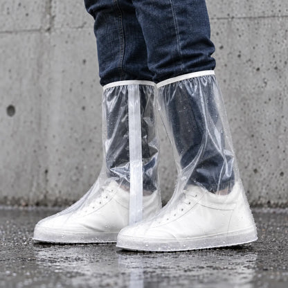 Clear knee-high overshoes with a white zip strip and raindrops, worn over white trainers against a grey concrete wall on wet ground. 3