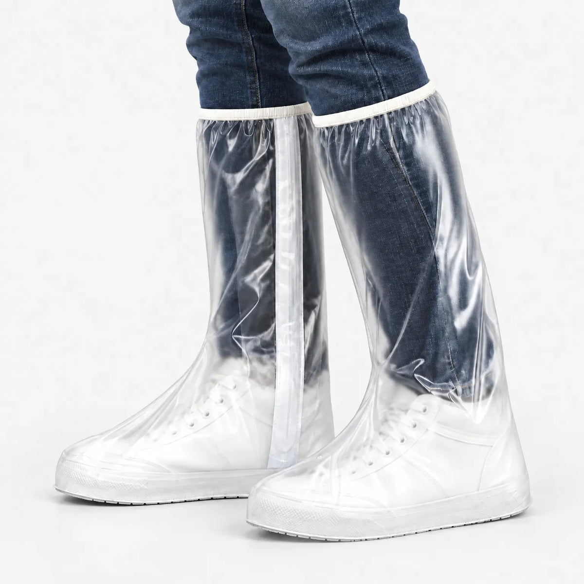 Studio side view of clear overshoes with white zip strip and trim, worn with jeans and white lace-up trainers on a plain background. 5