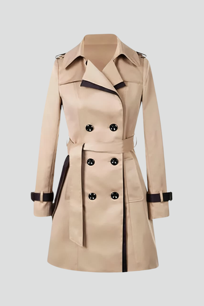 Beige double-breasted trench coat laid flat against a pale background, showing dark contrast trim, buttoned cuff straps and a tied belt. 1