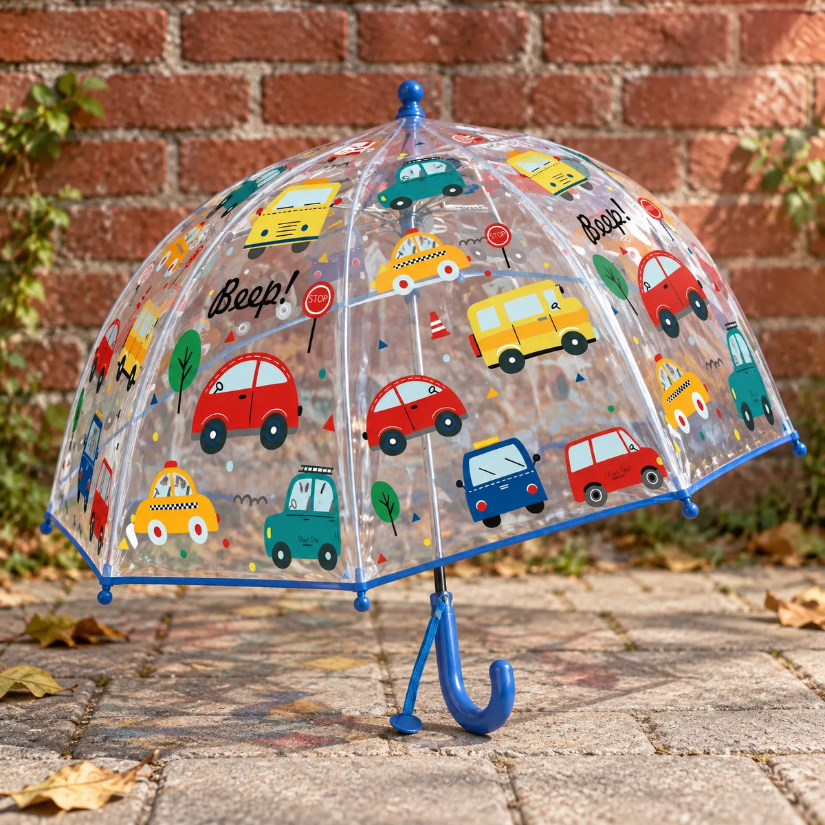 Clear dome umbrella printed with colourful cartoon cars, buses and taxis, blue trim and handle, standing on paving beside a brick wall. 3