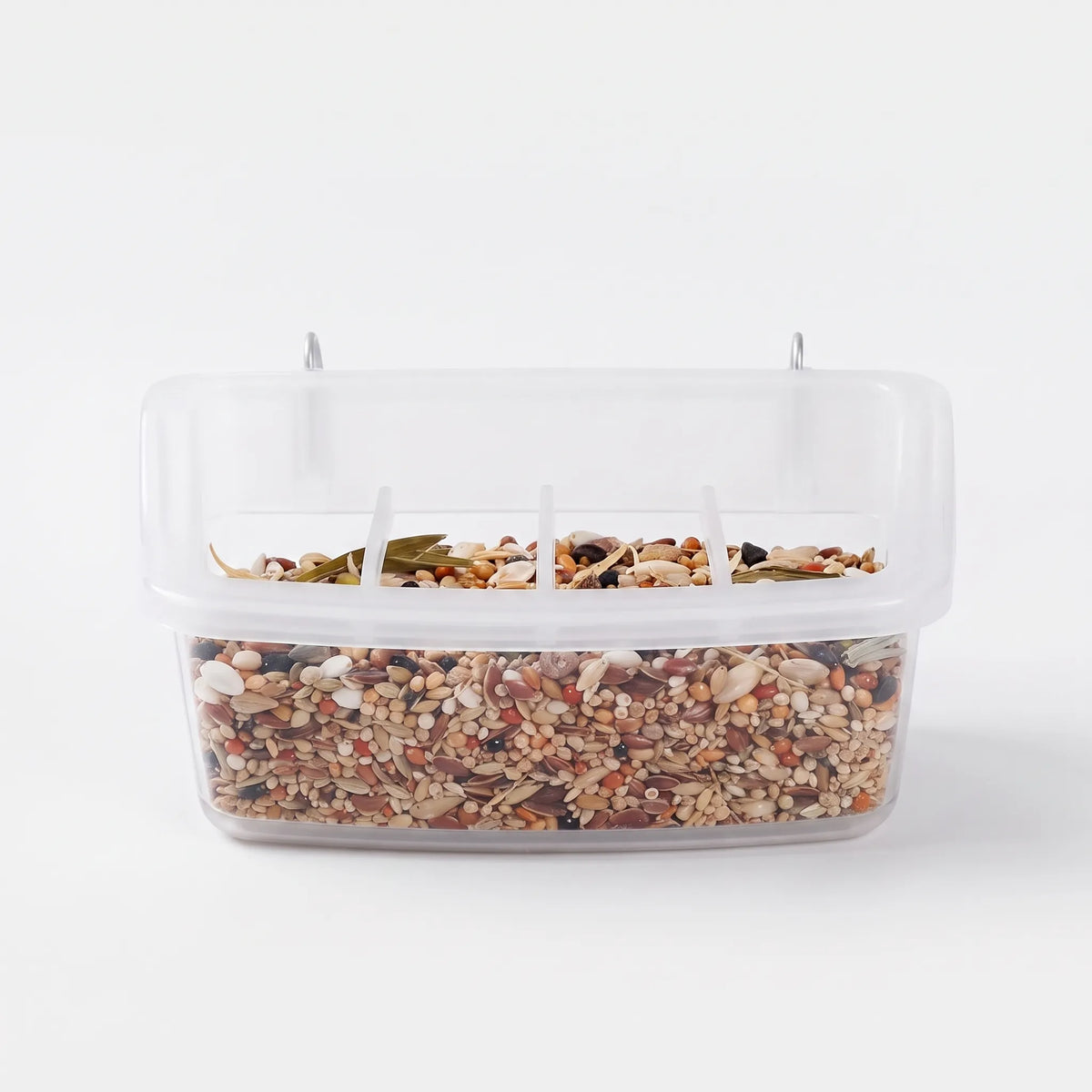 Front view of a clear plastic bird feeder filled with mixed seed, with metal hooks on top and divided feeding openings. 1