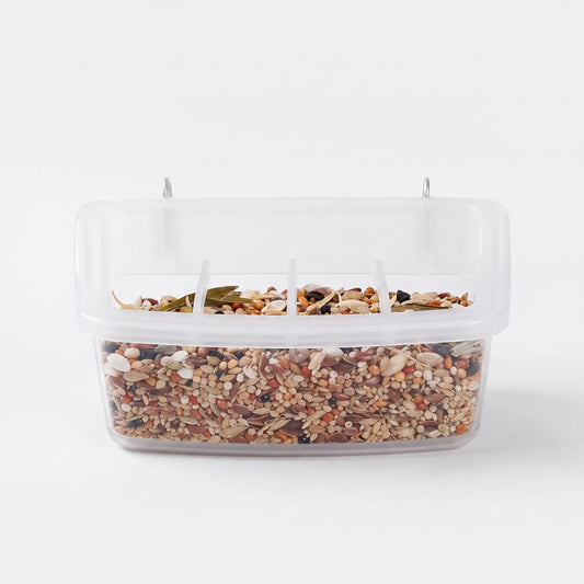 Front view of a clear plastic bird feeder filled with mixed seed, with metal hooks on top and divided feeding openings. 1