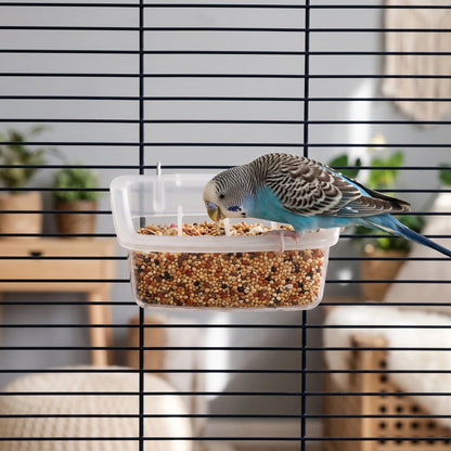 Blue and grey budgerigar perched on the feeder's rim and eating seed, with the feeder hung on a dark cage. 2