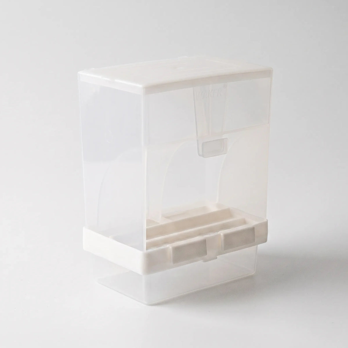 Three-quarter view from the other side of the empty dispenser, showing the lid, the clear seed chamber, white base clips and the tray below. 5