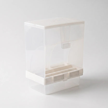 Three-quarter view from the other side of the empty dispenser, showing the lid, the clear seed chamber, white base clips and the tray below. 5