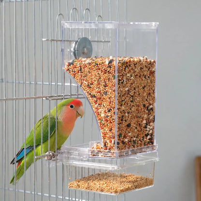 Green lovebird with a red face perched on the cage bars beside the seed-filled clear feeder, showing it in use. 2