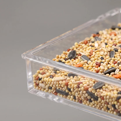 Close-up of the clear feeder's seed tray corner, showing mixed seeds and sunflower husks behind the transparent plastic wall. 3
