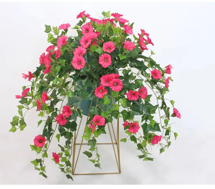 Pink flowers and trailing green foliage spilling over a dark pot on a gold geometric metal stand, shown against white. 3