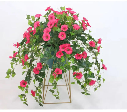 Pink flowers and trailing green foliage spilling over a dark pot on a gold geometric metal stand, shown against white. 3
