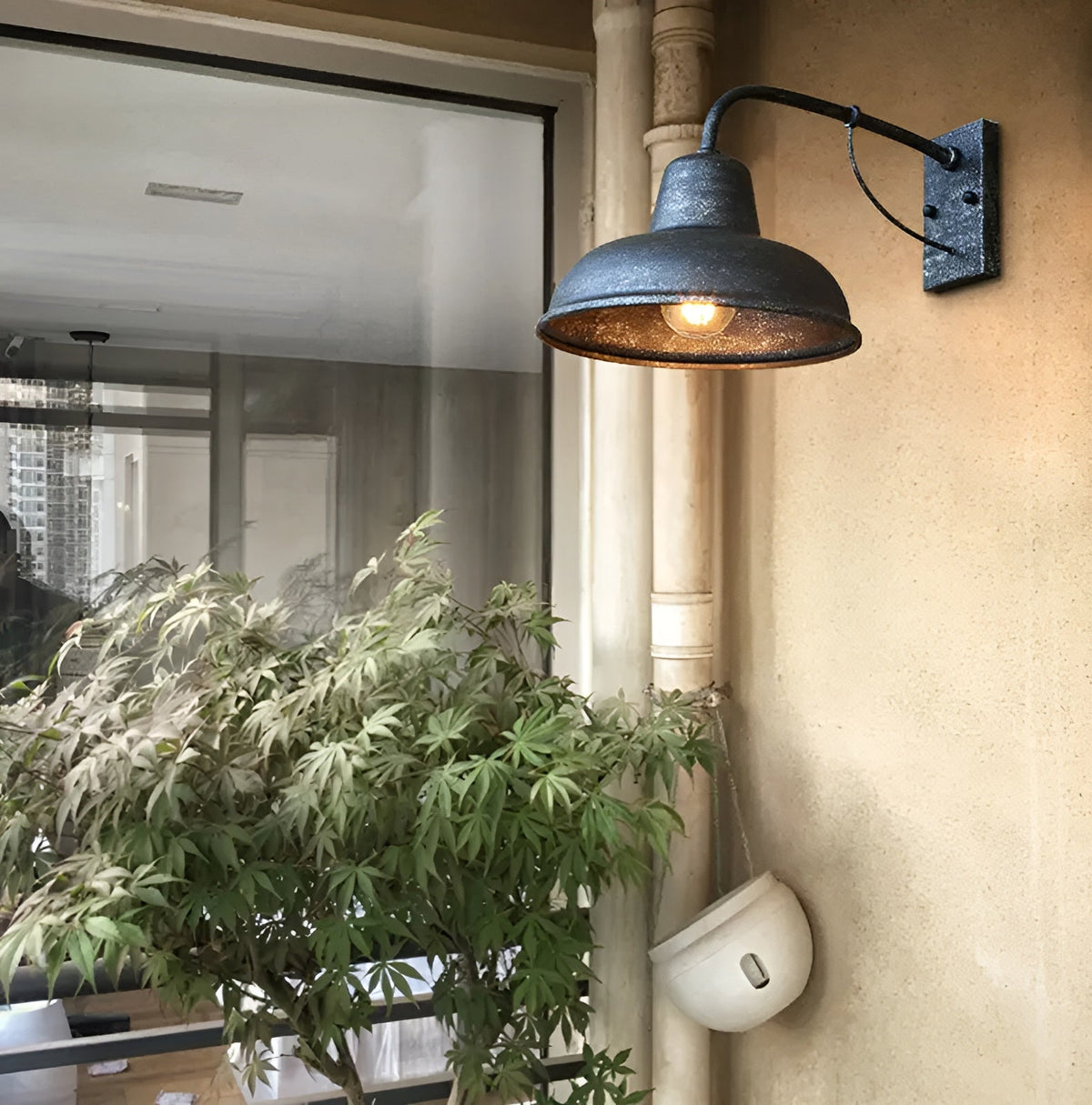 Lit wall light with speckled antique grey finish beside a window and a leafy potted plant on a sandy-coloured wall. 3