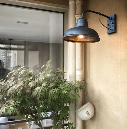 Lit wall light with speckled antique grey finish beside a window and a leafy potted plant on a sandy-coloured wall. 3