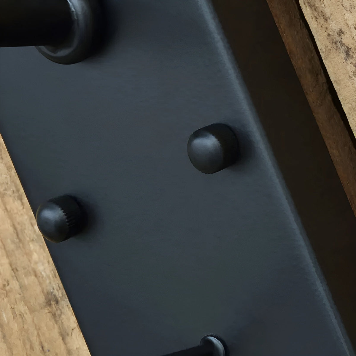 Close-up of the black rectangular backplate showing two rounded fixing studs and the arm joint, against rough wooden boards. 7
