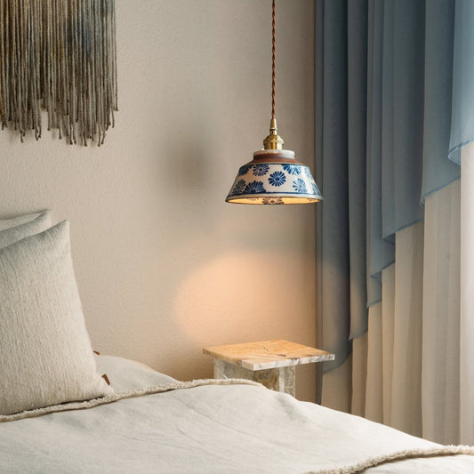 Blue floral ceramic pendant lit beside a bed, with cream bedding, a marble side table and soft blue curtains. 2